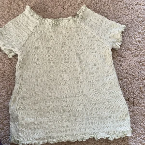 Ljusgrön smockad topp från H&M - Säljer en ljusgrön smockad topp från H&M med korta ärmar och vågig kant nertill. Toppen har en stretchig och figurnära passform, perfekt för en trendig look. Har en liten fläck på ryggen som knappt syns.