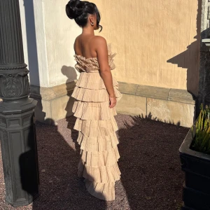 Beige volangklänning med axelbandslös design - Säljer en beige långklänning med flera lager volanger och axelbandslös modell. Klänningen har en elegant och unik look med plisserade detaljer och passar perfekt till speciella tillfällen.