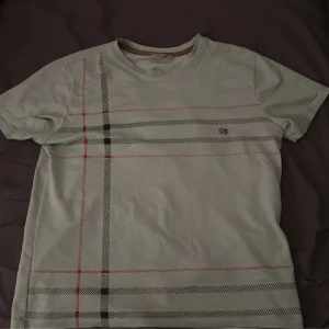 Ljusblå Tshirt från burberry - Säljer en vit t-shirt från burberry diskret rutigt mönster i svart, grått och rött på framsidan. T-shirten har rund halsringning och korta ärmar. det står M på den men den passar bara I S och XS