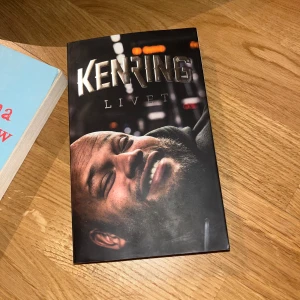 Ken Ring - Livet - Säljer boken 'Ken Ring: Livet' med ett omslag som visar Ken Ring leende mot kameran. Boken har svart bakgrund och titeln är tryckt i stora, silvriga bokstäver. Perfekt för dig som gillar biografier och musik.