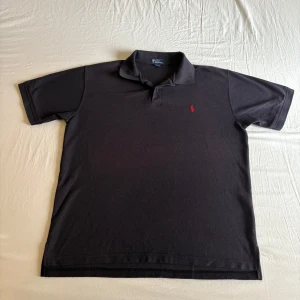 Svart pikétröja – Ralph Lauren  - Stilren klassiker du alltid kan lita på! Svart pikétröja från Ralph Lauren med den ikoniska röda loggan på bröstet. Perfekt passform, tidlös design och funkar lika bra till jeans som till chinos. Storlek: L Skick: Mycket bra 