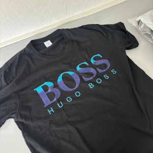 Svart t-shirt från Hugo Boss - Snygg svart t-shirt från Hugo Boss med stor logga i blått och lila framtill. Klassisk rund halsringning och normal passform. Perfekt för dig som gillar stilrena och trendiga plagg med tydlig branding.NySKICK! STRL XL. Passar större varianten av L
