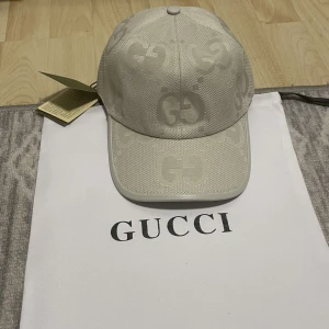 Beige keps från Gucci med GG-mönster - Snygg beige keps från Gucci med diskret GG-logga i mönstret. Kepsen har böjd skärm och klassisk passform. Perfekt accessoar för en stilren look.