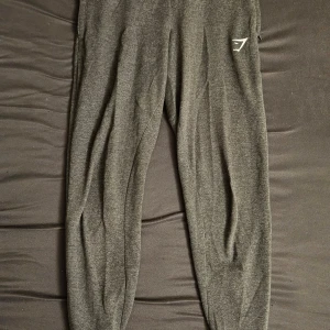 Mörkgrå träningstights från Gymshark - Säljer ett par mörkgrå mjukisbyxor från Gymshark i storlek XS. Byxorna har en diskret logga på låret och dragkedja vid benslutet. Perfekta för gymmet eller löpning.