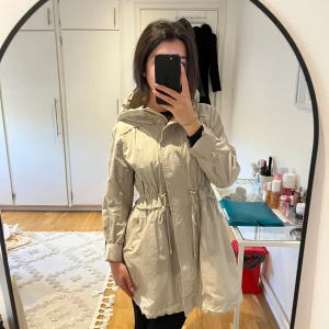 Beige tunn vårjacka med huva och dragsko - Säljer en beige parkajacka med huva och dragsko i midjan. Jackan har guldiga detaljer, långa ärmar och stängs med dragkedja och knappar framtill. Perfekt för övergångssäsonger och har en avslappnad passform. Pris går att diskuteras ✨