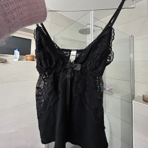 Svart spetslinne med rosett - Säljer ett svart linne med spetsdetaljer och en liten rosett framtill. Linnet har smala axelband och är tillverkat i ett tunt, genombrutet material som ger en elegant look.
