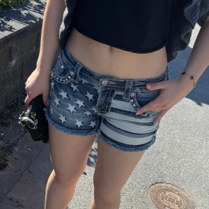 Jeansshorts med stjärnor och ränder - Säljer ett par coola jeansshorts från Miss Me med amerikanskt flaggmönster – stjärnor på ena sidan och ränder på andra. Shorts med låg midja, slitna detaljer, nitar och råa kanter. Perfekta för sommaren!