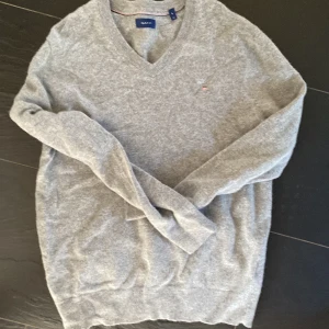 Grå v-ringad tröja från Gant - Säljer en klassisk grå v-ringad tröja från Gant med broderad logga på bröstet. Tröjan är i mjukt material och har långa ärmar samt ribbade muddar vid ärmslut och nederkant. Perfekt för lager-på-lager-stil. Passar mer som M då den krympt lite
