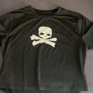 Svart t-shirt med dödskalletryck - Cool svart t-shirt med ett stort vitt dödskalle- och korsade ben-tryck på bröstet. T-shirten har korta ärmar och en klassisk rund halsringning. Perfekt för dig som gillar edgy och stilrena plagg. Plagget är droppad och jag vet inte vart jag köpte den. Aldrig använd.