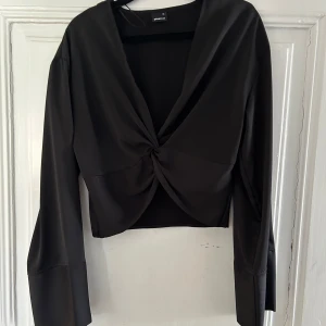 Svart croppad topp med knut från Gina Tricot - Säljer en svart croppad topp från Gina Tricot i storlek XS. Toppen har lång ärm, djup v-ringning och en snygg knutdetalj framtill. 