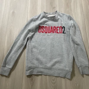 Grå sweatshirt från Dsquared2 - Säljer en stilren grå sweatshirt från Dsquared2 med logga i rött och svart broderi på bröstet. Tröjan har rund halsringning och långa ärmar. Perfekt för en avslappnad och trendig look.