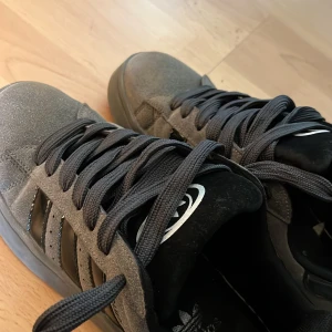 Grå Adidas Campus sneakers - Säljer ett par grå Adidas Campus sneakers med svarta ränder och svarta skosnören. Skorna har en klassisk låg profil och rund tå. Perfekta för dig som gillar stilrena och bekväma sneakers.