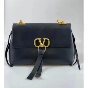 Svart axelväska från Valentino - Snygg svart axelväska från Valentino med guldiga metalldetaljer och stort V-logga framtill. Väskan är i skinn och har ett innerfack med dragkedja samt justerbar axelrem. Perfekt för att lyfta din outfit med en lyxig touch.