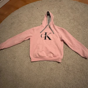 Rosa hoodie med CK-tryck från Roly - Mysig rosa hoodie med stor Calvin Klein logga på bröstet. Tröjan har huva med grå insida och dragsnören samt en stor magficka framtill. 