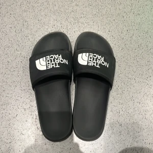 Svarta badsandaler från The North Face - Svarta sandaler från The North Face med vit logga på remmen. Klassisk slip-on modell med platt sula och öppen tå, perfekt för stranden eller poolen. Snygg och enkel design som passar till det mesta.