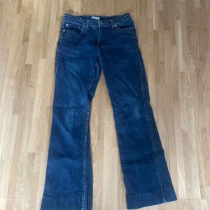 Mörkblå bootcut jeans - Säljer ett par mörkblå jeans med bootcut passform och klassiska fem fickor. Jeansen har normalhög midja, knapp- och dragkedjestängning samt dekorativ etikett bak i midjan. Perfekta för dig som gillar en lite vidare benvidd.