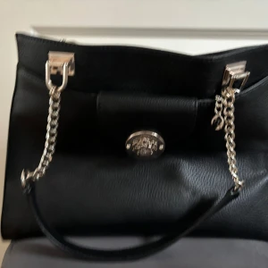 Svart handväska med kedjedetaljer från Guess - Snygg svart handväska från Guess med silverfärgade kedjor och detaljer. Väskan har en klassisk rektangulär form, dragkedja på baksidan och logga framtill. 