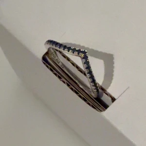 Silvrig ring med mörkblåa stenar från Pandora - Säljer en V formad silverring från Pandora. Ringen har små mörkblåa stenar längs ovansidan och har stämpeln 925 ALE 54 på insidan. Storlek 54 (alltså 7). Inga tecken på användning.