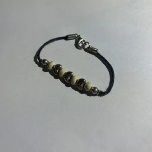 Svart armband med pärlor - Snyggt armband med svart snöre och pärlor i silver och vitt. Pärlorna har blommönster och armbandet stängs med en metallhake. Perfekt accessoar för att lyfta din stil. Köpte det i Sydafrika 🤍