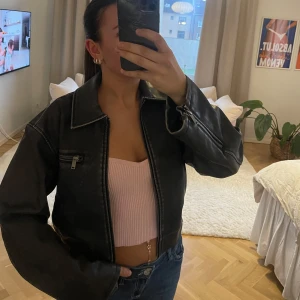 Svart skinnjacka med dragkedja - Säljer en svart skinnjacka med dragkedja och bred krage. Jackan har en ficka med dragkedja på bröstet och markerade sömmar. Passformen är normal och jackan har långa ärmar. Perfekt för en cool och stilren look.