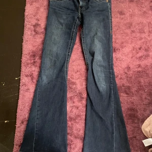 Mörkblå bootcut jeans från True Religion - Säljer ett par mörkblå jeans från True Religion med bootcut och lågmidjad passform. Jeansen har klassiska kontrastsömmar, ficklock med knapp bak och detaljerad bakficka. Perfekta för dig som gillar en tidlös jeansstil.