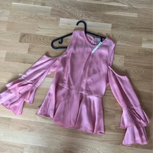 Rosa satinblus med volangärmar - Säljer en superfin rosa blus i satin med trendiga volangärmar och öppna axlar. Blusen har en lös passform och är perfekt för dig som vill sticka ut lite extra. Supersnygg till både jeans och kjol!