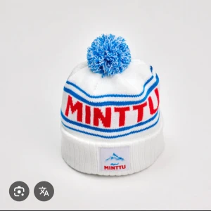 Vit och blå mössa med tofs från Minttu - Vit mössa med blå och röda detaljer samt blå tofs på toppen. Minttu-logga framtill och texten 'MINTTU' i rött runt om. Klassisk retrostil med ribbad nederkant och mjukt material.