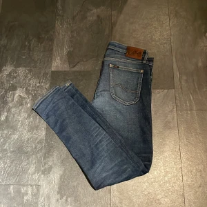 Lee jeans - Säljer ett par klassiska blå jeans från Lee med fem fickor och läderpatch bak i midjan. Jeansen har en rak passform och subtila slitningar framtill för en snygg, vardaglig look.
