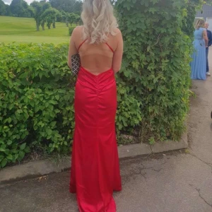 Röd balklänning med öppen rygg - Säljer en elegant röd långklänning med smala axelband och öppen rygg. Klänningen har en figurnära passform och är tillverkad i ett glansigt material som ger en lyxig känsla. Perfekt för bal eller festliga tillfällen.