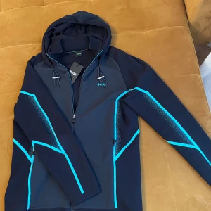 Boss jacka - Säljer en svart hoodiejacka från BOSS med dragkedja och huva. Jackan har neon blåa detaljer längs ärmar och sidor samt BOSS-loggan på bröstet. Rekommenderar den till dig som gillar en sportig samt bekväm stil💯                                        Säljs pågrund av att jag inte använder den längre då jag har köpt nya jackor, jackan är verkligen felfri exakt som alla mina andra varor💯🫡