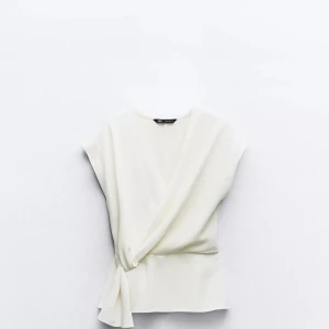 Vit omlottopp med knyt - Säljer en stilren vit omlottopp från Zara med knyt i sidan. Toppen har korta ärmar och en elegant v-ringning. Perfekt för dig som gillar en enkel men snygg look.