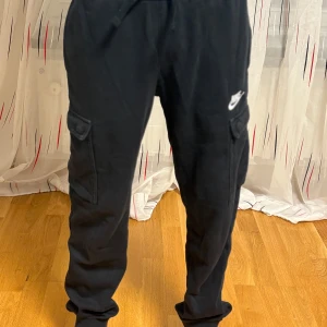 Svarta cargobyxor från Nike - Svarta cargobyxor från Nike med dragsko i midjan och resår vid bensluten. Byxorna har sidofickor med lock och en vit Nike-logga på vänster lår. Perfekta för en avslappnad och sportig stil. 