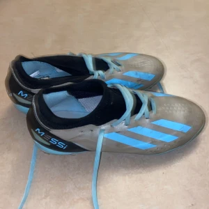 Adidas Messi fotbollsskor med blå detaljer - Säljer ett par Adidas Messi fotbollsskor i grått med ljusblå ränder och detaljer. Skorna har snörning och låg profil, perfekt för dig som vill sticka ut på planen. Messi-logga på hälen och klassisk Adidas-design. Och swisha numret 0769313560