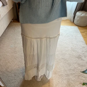 Vit långkjol med spetsdetaljer - Säljer en vit långkjol med fina spetsdetaljer och lätt rynkad struktur. Kjolen har en bohemisk känsla och är perfekt för varma dagar. Resår i midjan för bekväm passform.