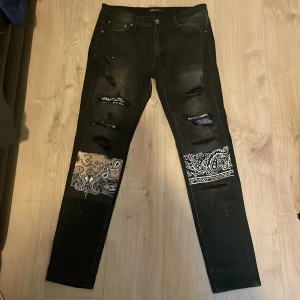 Svarta jeans från med bandanamönster och slitningar från Hesntls - Säljer ett par svarta jeans från Hesntls med vita bandanamönster på ben och bakficka. Perfekta för dig som vill sticka ut med detaljer och streetstyle.