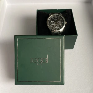 Silverfärgad klocka från Regal - Stilren klocka från Regal med svart urtavla och silverfärgat armband i metall. Klockan har flera små urtavlor och romerska siffror. Levereras i original grön ask med Regal-logga.(OBS!! Minutvisaren funkar inte men går antagligen att fixa) 