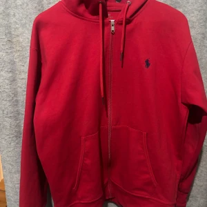 Röd hoodie från Polo Ralph Lauren - Säljer en klassisk röd hoodie från Polo Ralph Lauren med dragkedja och två fickor framtill. Tröjan har huva med dragsko och en broderad blå logga på bröstet. Perfekt för en avslappnad stil.