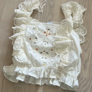 Vit broderad blus med spets och volang - Superfin vit blus från Zara med broderade blommor framtill, spetsdetaljer och volanger längs axelbanden. Blusen har en croppad passform och är tillverkad i bomull. Perfekt för sommardagar!