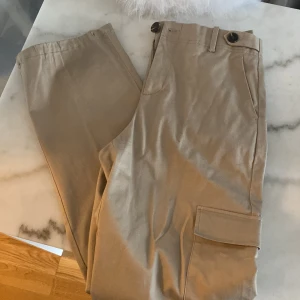 Beige cargopants med fickor - Snygga beige cargopants med stora fickor på sidorna och klassisk passform. Byxorna har bälteshällor och stängs med knapp och dragkedja. Perfekta för en avslappnad och trendig stil.