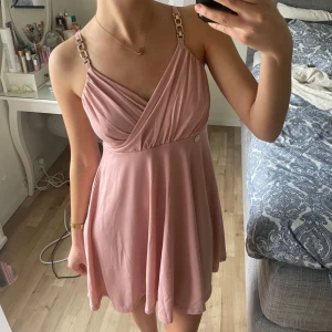 Ljusrosa omlottklänning med kedjedetaljer - Säljer en söt ljusrosa kortklänning med omlottliv och smala axelband med kedjedetaljer. Klänningen har en lätt utställd kjol och v-ringning framtill. Perfekt för sommarens tillfällen!