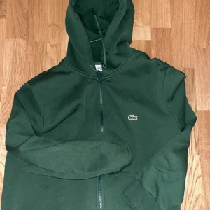 Mörkgrön hoodie från Lacoste - Säljer en mörkgrön hoodie från Lacoste med dragkedja och klassisk krokodillogga på bröstet. Tröjan har huva och långa ärmar, perfekt för en avslappnad stil. Classic fit-modell.