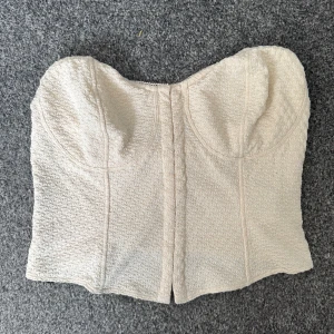 Beige tubtopp från H&M Divided - Säljer en beige tubtopp från H&M Divided med snyggt strukturerat mönster i tyget. Toppen är axelbandslös och har en lätt formad byst för extra stöd. Perfekt att styla till olika outfits!
