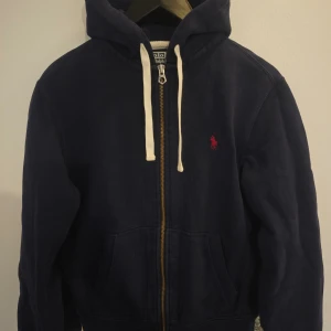 Mörkblå hoodie från Polo Ralph Lauren - Säljer en snygg mörkblå hoodie från Polo Ralph Lauren med dragkedja och broderad logga i rött. Den har en justerbar huva med vita snören och är tillverkad i en blandning av bomull och polyester. Perfekt för en avslappnad stil.