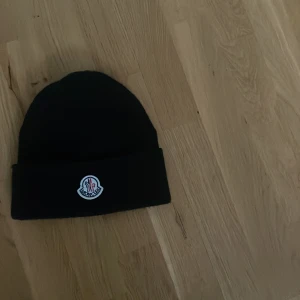 Svart mössa från Moncler - Snygg svart mössa från Moncler med broderad logotyp framtill. Tillverkad i 100% akryl, perfekt för kyliga dagar. Stilren design som passar till det mesta.