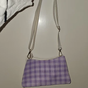 Lila rutig axelväska - Snygg lila axelväska med ett klassiskt rutigt mönster. Väskan har en justerbar axelrem i vitt och en praktisk dragkedja upptill. Perfekt för att ge din outfit en färgklick!