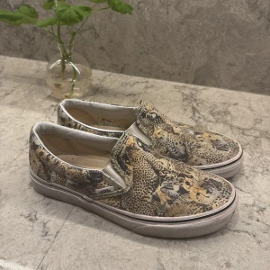 Leopardmönstrade slip-on sneakers från Vans - Snygga slip-on sneakers från Vans med ett coolt leopardmönster. Skorna har en bekväm passform och är perfekta för en avslappnad stil. De har en vit sula som kontrasterar snyggt mot det djärva mönstret.