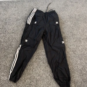 Svarta träningsbyxor från Adidas - Snygga svarta träningsbyxor från Adidas med de klassiska tre vita ränderna längs sidorna. Byxorna har elastisk midja med snörning och resår vid bensluten. Perfekta för träning eller en avslappnad stil. Adidas-loggan syns på flera ställen för en sportig look.