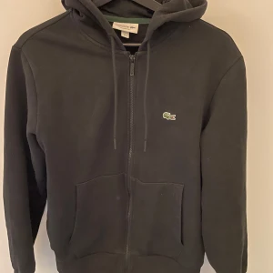 Svart Lacoste hoodie - Säljer en klassisk svart hoodie från Lacoste med dragkedja och huva. Tröjan har långa ärmar och en broderad logotyp på bröstet. Perfekt för en avslappnad stil. Storleken är M men skulle säga att den också passar för S.