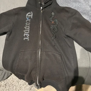 Made by Trappers hoodie svart - Säljer en svart hoodie med dragkedja och coolt ormmotiv på bröstet. Texten 'Trapper' i glittriga bokstäver ger en extra edge. Perfekt för en avslappnad stil.