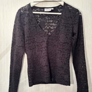 Svart spetsblus från Kappahl - Elegant svart spetsblus från KappAhl med v-ringning och långa ärmar. Perfekt för en stilren look med ett vackert spetsmönster som ger en sofistikerad touch. Passar utmärkt till både vardag och fest.